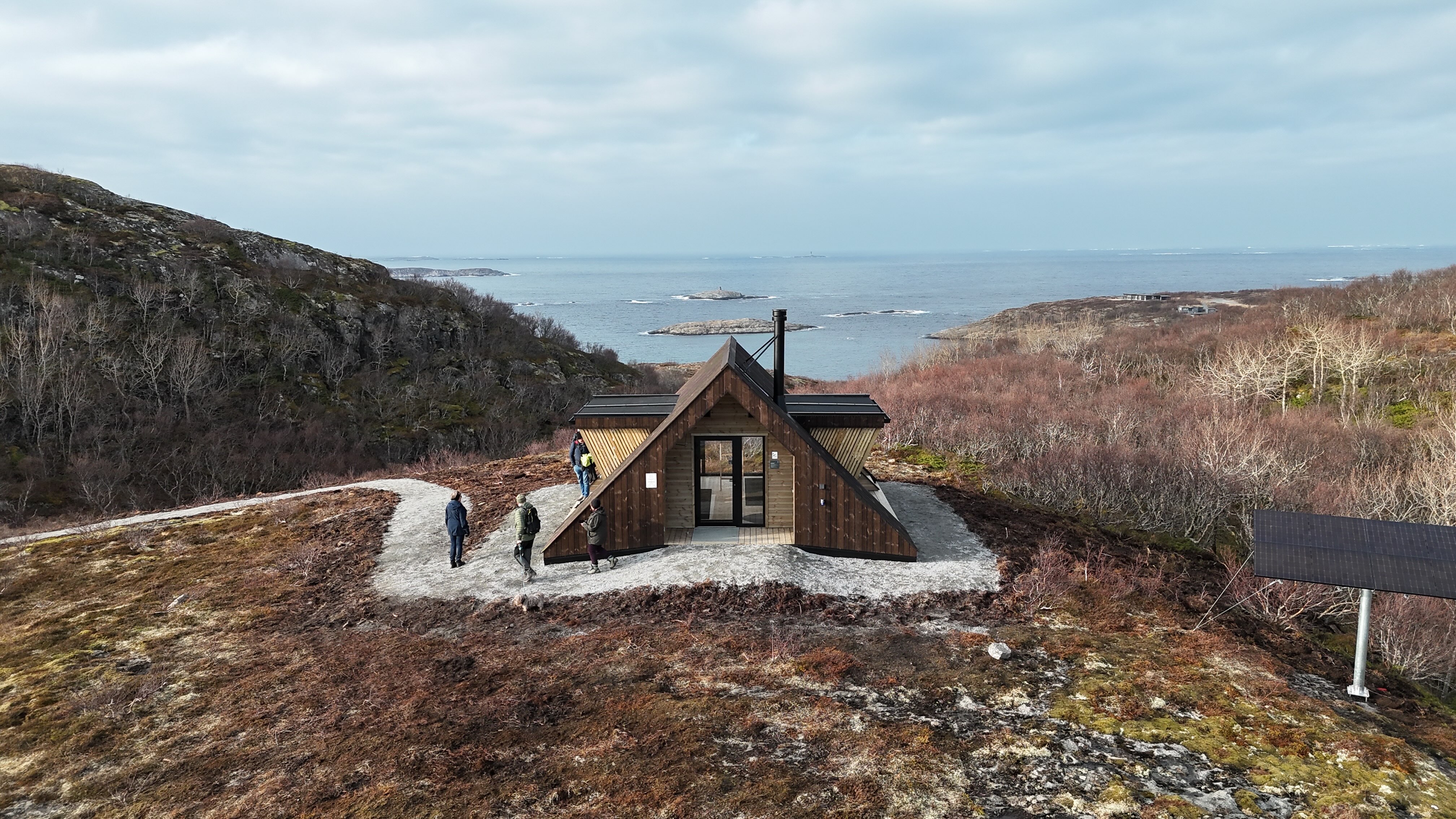 Dagsturhytta ved Utrosjøen ligger på en åpen høyde med utsikt mot hav og holmer, omgitt av karrig kystlandskap, med flere turgåere på stien og et solcellepanel like ved. Foto: Jonas Ali Ghanizadeh