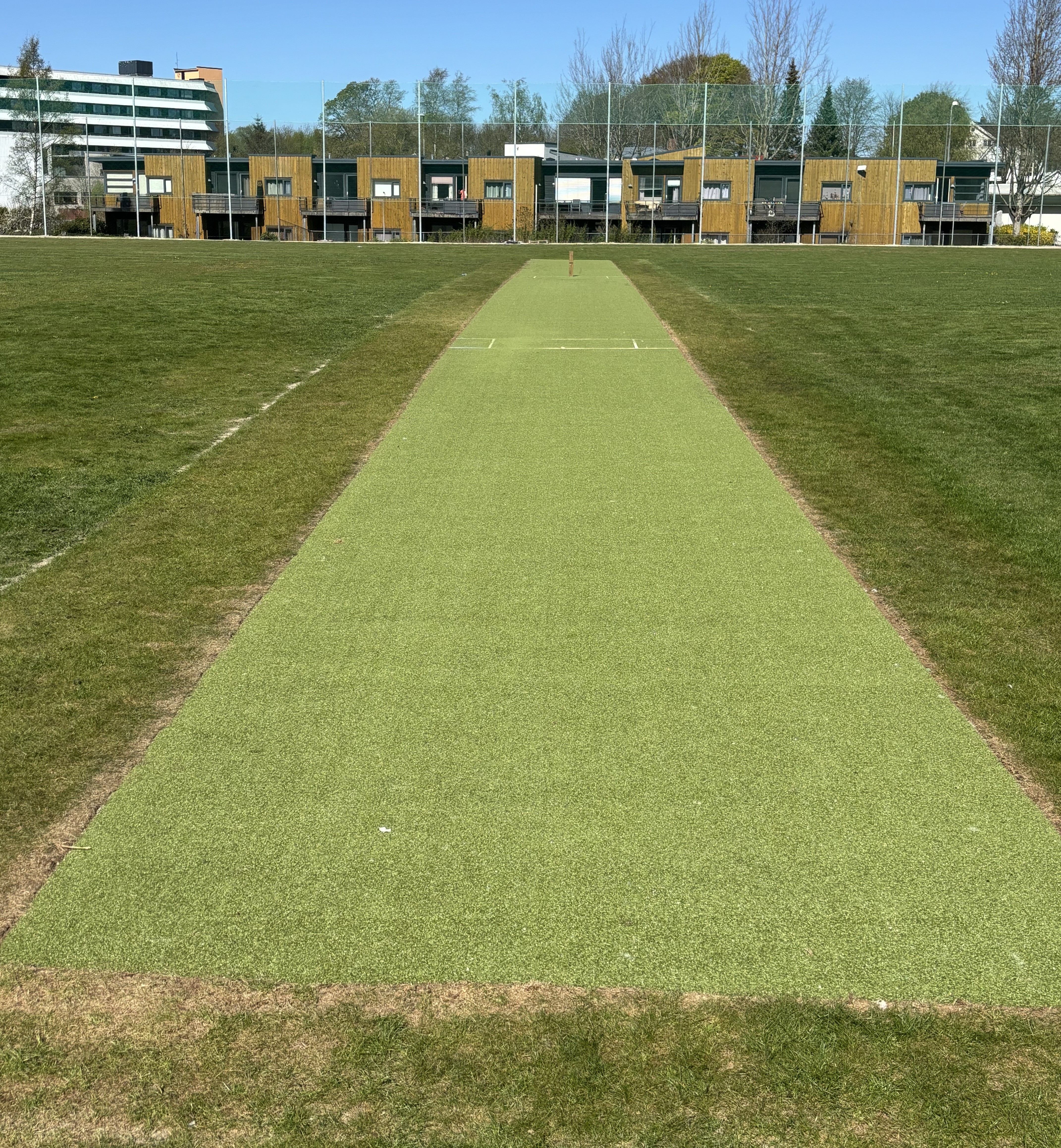 Bilde viser cricketbanen av kunstdekke som ligger midt på en gressbane, med bygninger og nettgjerde i bakgrunnen.
