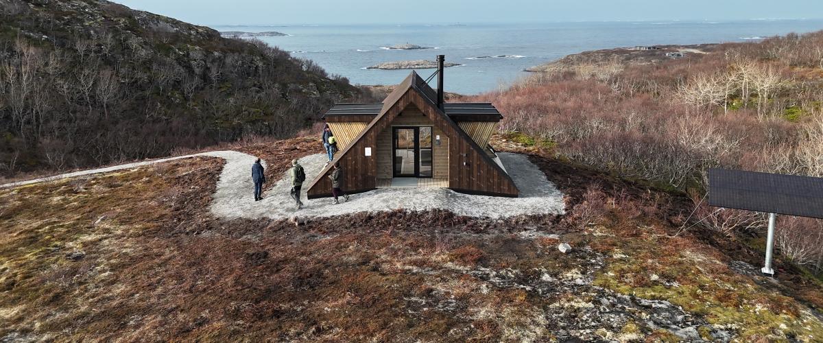 Dagsturhytta ved Utrosjøen ligger fritt til på høydedraget over kystlandskapet i Åfjord, med tursti, solcellepanel og vid utsikt mot havet og de nakne holmene utenfor. Dagsturhytta ved Utrosjøen ligger på en åpen høyde med utsikt mot hav og holmer, omgitt av karrig kystlandskap, med flere turgåere på stien og et solcellepanel like ved. Foto: Jonas Ali Ghanizadeh