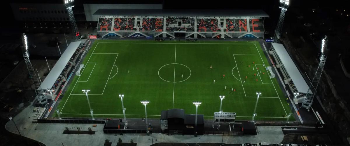 Bildet viser Åsane Arena opplyst med flomlys under en kveldskamp. Tribunene er delvis fylt, og banen er klart belyst, med spillere i aksjon.