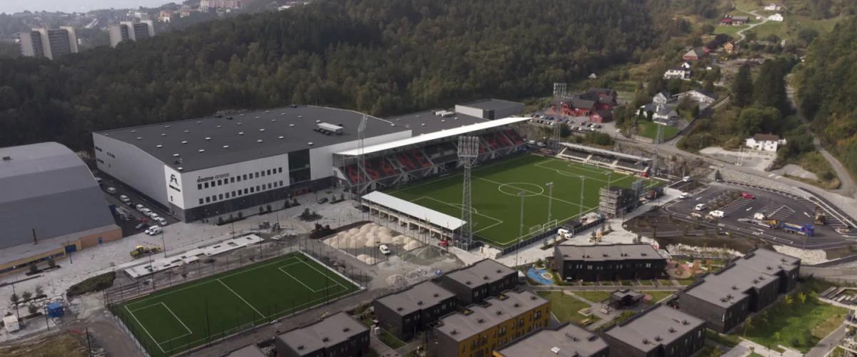 Bildet viser en oversikt over Åsane Arena, et idrettsanlegg med flere fotballbaner, tribuner og bygninger. Arenaen ligger omgitt av skog og boligområder, med fjell og åser i bakgrunnen. Anlegget har både utendørs og innendørs fasiliteter, inkludert en hovedbane med tribuner og flomlys.