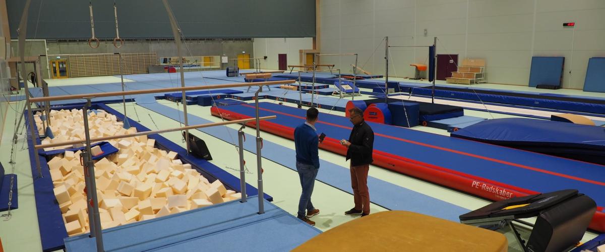 TIL LEIE: Åsane trengte en turnhall - og turnhall fikk de! Den kan leies av bergenske idrettslag, helt kostnadsfritt. Grunnen til det? Gratisprinsippet til Bergen kommune. Foto: Sørli Arkitekter Bildet viser turnhallen på Åsane Arena med diverse turnutstyr som ringer, bommer, trampoliner og skumgroper. To personer står og snakker i hallen.