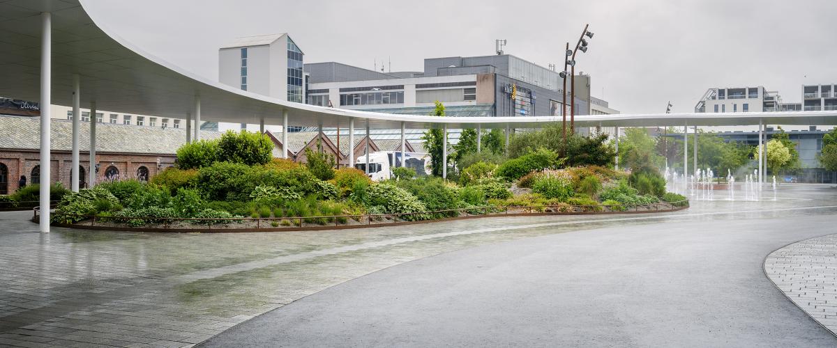 Den hvite ringen Lysningen omkranser Ruten bypark. Foto: Arkitektbedriftene/Kim Ramberghaug. Utsikt under den hvite, buede ringen på Ruten, med gamle murbygninger med buede vinduer og et beplantet bed i midten.