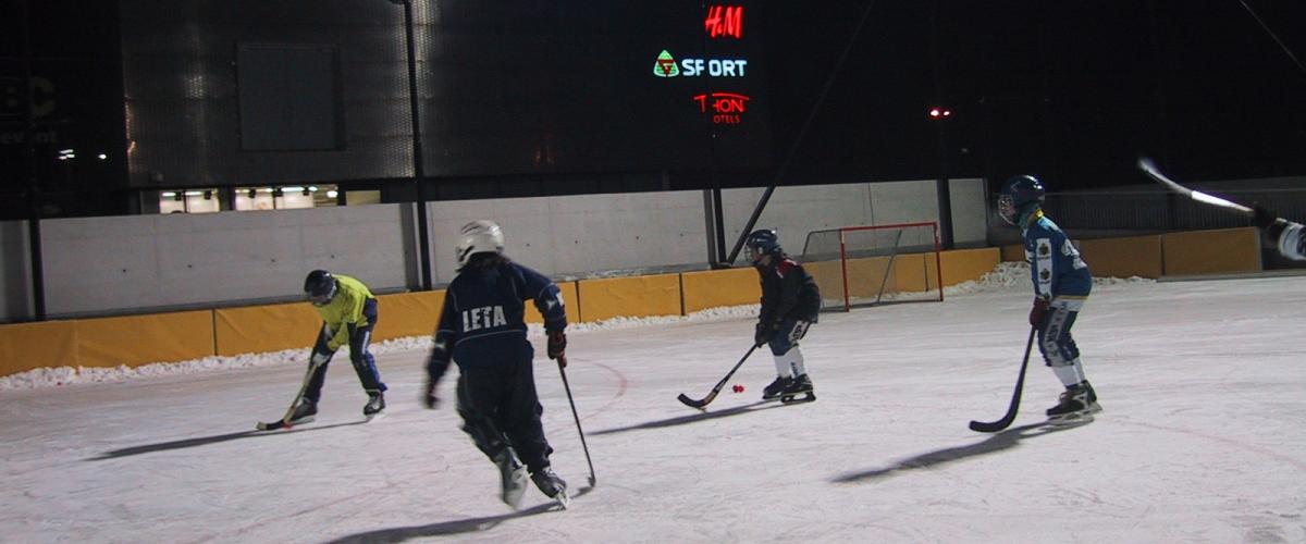 Barn som spiller bandy på Bergbanen på kveldstid. I bakgrunnen ser man Ullevaal Stadion Kjøpesenter.