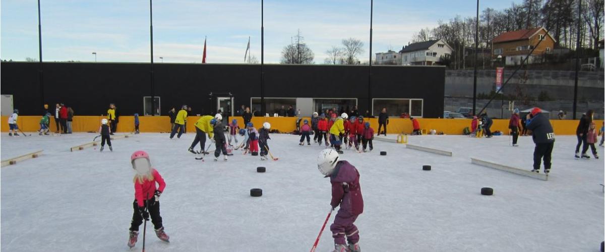 Jenter som spiller bandy på bergbanen. I bakgrunnen ser man klubbhuset som tilhører bandybanen. Blå himmel.