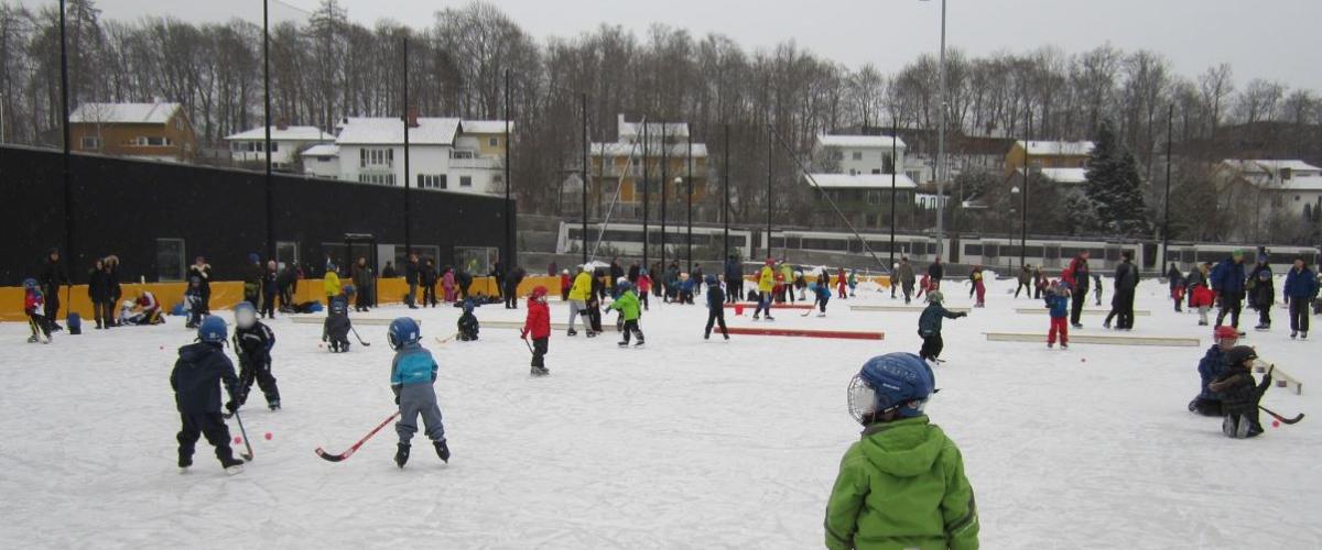 Gutter som spiller bandy på bergbanen. I bakgrunnen ser man klubbhuser og boligfelt.En grå dag. 