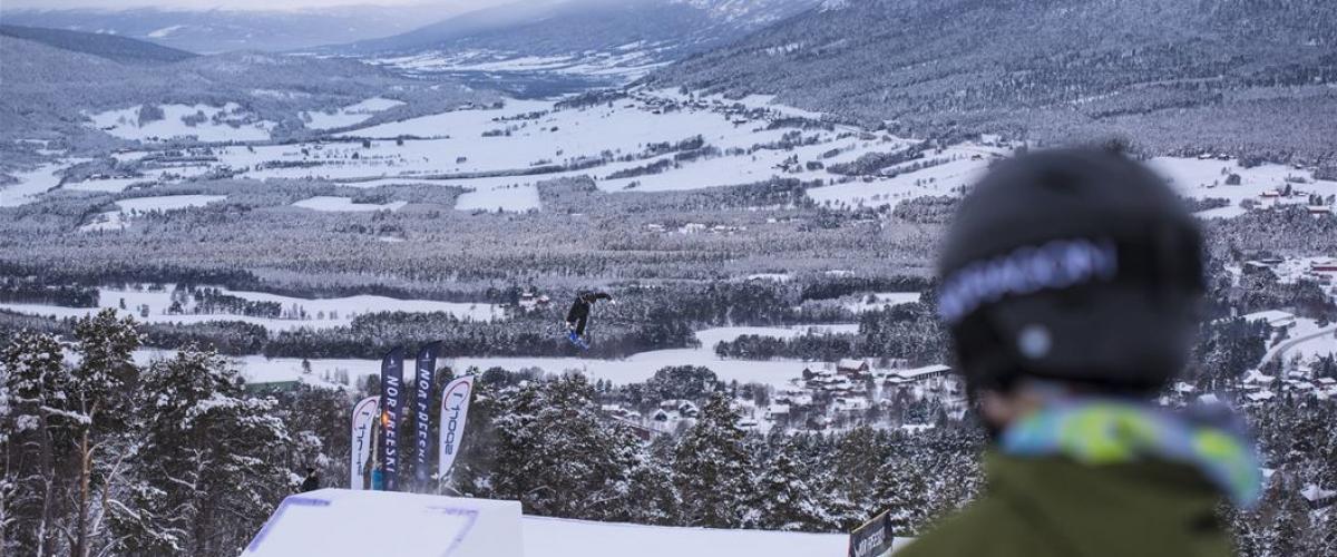 Bilde av en skikjører som ser på et stort hopp, der en annen skikjører nettopp har lettet, og har fått godt med luft under seg. 