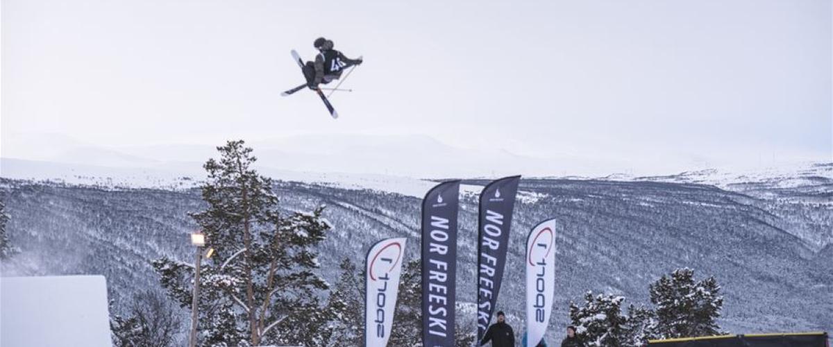 Bilde av en skikjører som nettopp har forlatt hoppkanten, og som svever høyt i lufta. Fjell og åser i bakgrunnen. 