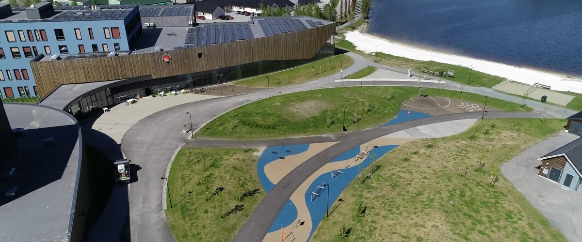 Stort idrettsbygg og badeland med park, strand og fjord utenfor.