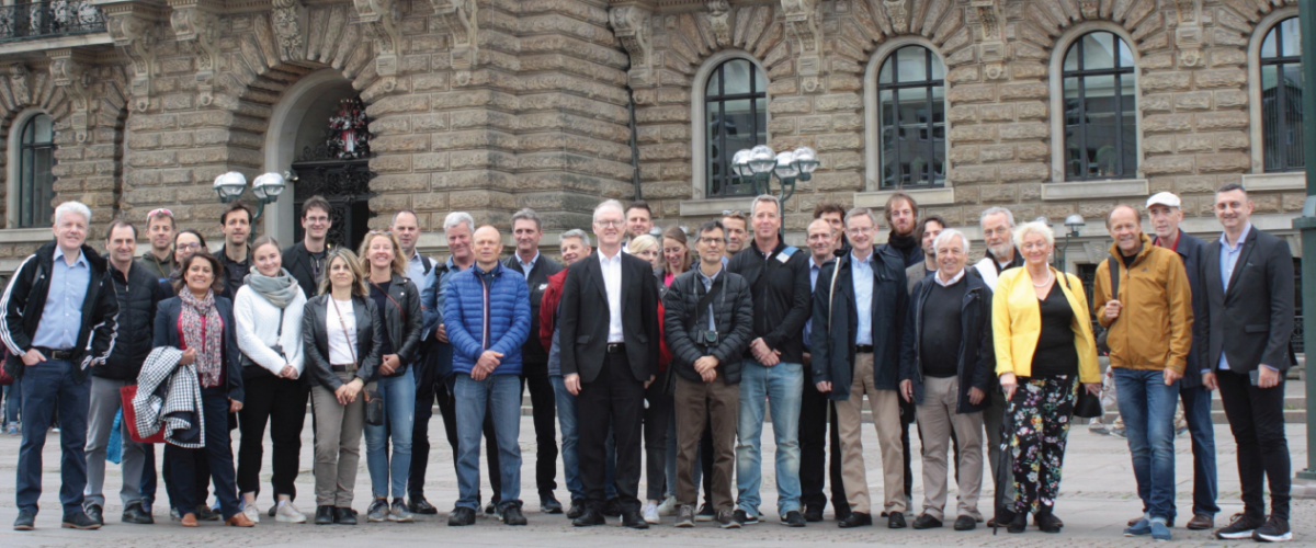 Gruppe med mennesker poserer smilende foran stort murbygg. 