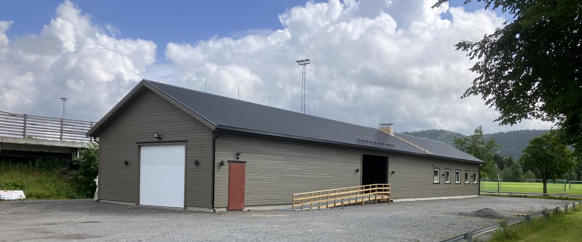 Fasade klubbhus fra annen vinkel. Fra denne vinkelen ser man garasjeport på den motsatte kortsiden av bygget. Man ser også en tilpasset rampe som leder inn til hovedinngangen midt på langsiden av bygget. Gruslagt plass rundt bygget, med fotballbane og skog i bakgrunnen.