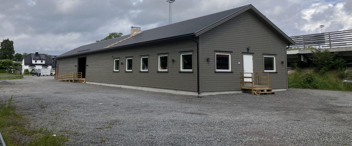 Fasade klubbhus. Bygget har grå kledning, sorte takplater,  syv vinduer i den ene enden av bygget. Gruslagt område rundt, med bolighus og fotballbaner i bakgrunnen.