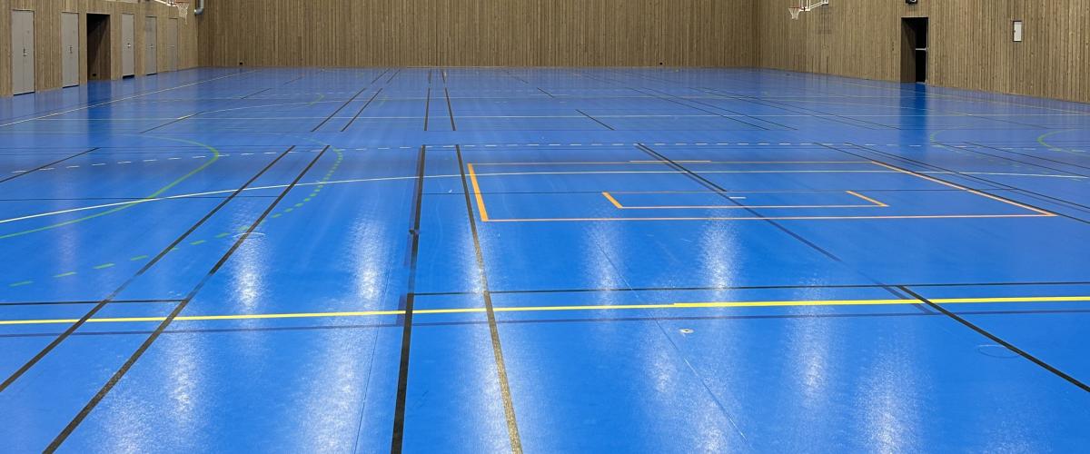 Idrettshallen har blått gulv med markering for blant annet badminton. Veggene er dekket med trespiler. 