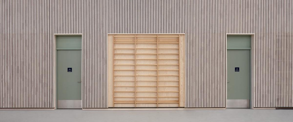 Vegg i idrettshall med ribbevegg i midten og to grønne dører på hver side, begge merket med rullestolsymbol. Veggen er kledd i stående trespiler, og hallgulvet har fargede banemarkeringer.