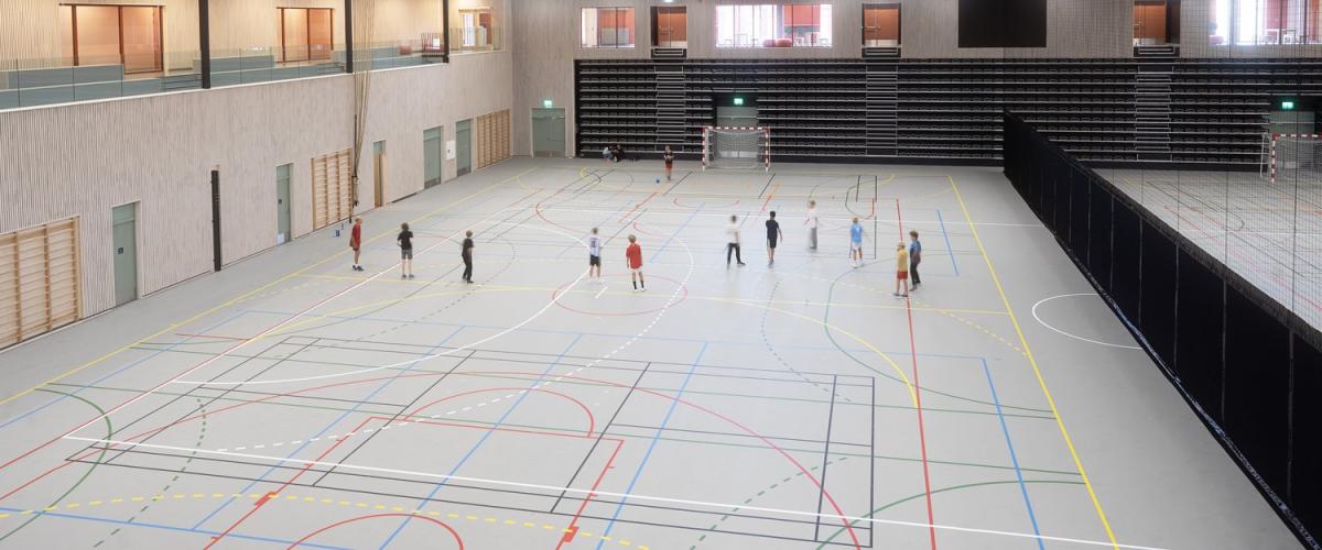 Oversiktsbilde av stor idrettshall med flere håndballbaner og elever i aktivitet. Hallen har teleskoptribune, høyt svart tak med synlige ventilasjonsrør, og store vinduer inn mot møterom og publikumsarealer.