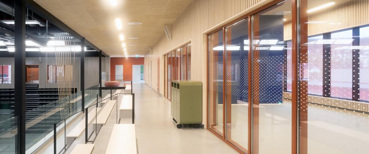 Innendørs tribuneområde med benkeseter og glassrekkverk i en idrettshall. Til høyre er det et innglasset møterom med oransje vegger og prikket folie på vinduene. Taket er dekket med himlingsplater i treullsement.