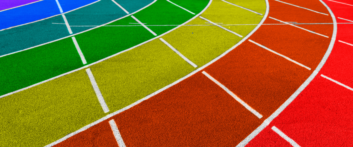 Bildet viser en løpebane på en idrettsplass malt i regnbuens farger. Banen har flere spor i fargene lilla, blå, grønn, gul, oransje og rød, som symboliserer mangfold og inkludering i forbindelse med Pride-bevegelsen. De hvite linjene som skiller sporene er tydelige mot de sterke fargene.