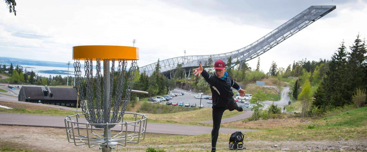 Anlegget er mye brukt av frisbeekastere på alle nivåer, og ligger enkelt til med tanke på kollektivtransport. Foto: Skiforeningen. Person som kaster frisbee mot en av anleggets kurver. Hoppbakken er i bakgrunnen.