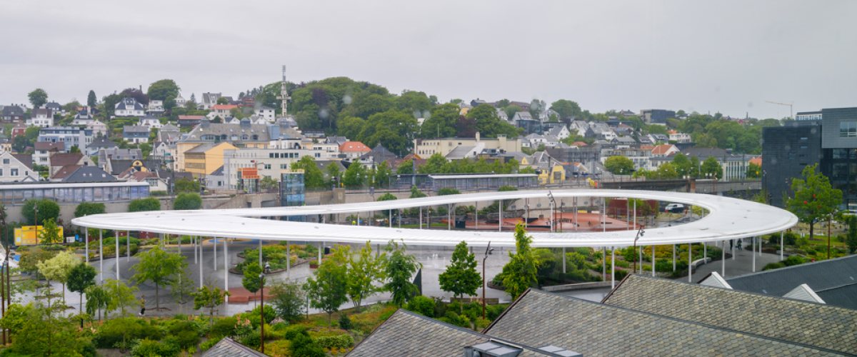 Ruten bypark sett ovenfra, med Lysningen og skateparken i midten. Foto: Arkitektbedriftene/Kim Ramberghaug. Luftfoto av Ruten bypark som viser den ringformede, hvite takkonstruksjonen Lysningen sett ovenfra, med skateparken inni ringen og Sandnes sentrum og boligbebyggelse i bakgrunnen.