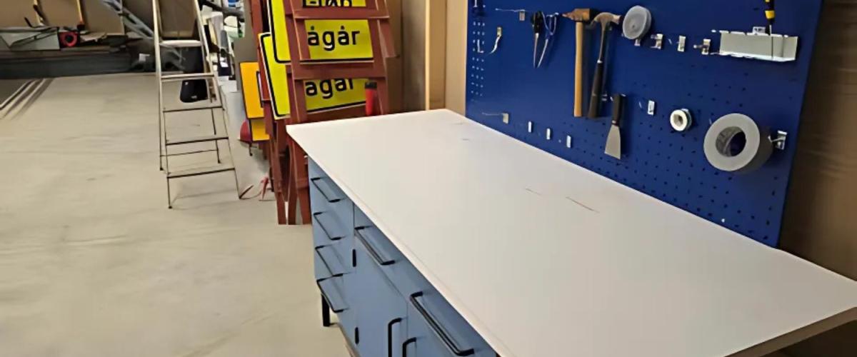 Bilde av arbeidsbenk i garasje/lager/verksted. Over arbeidsbenken står en verktøytavle med diverse håndverktøy og hyller fylt med utstyr og skilt. En stige og flere sperreskilt står langs veggen.