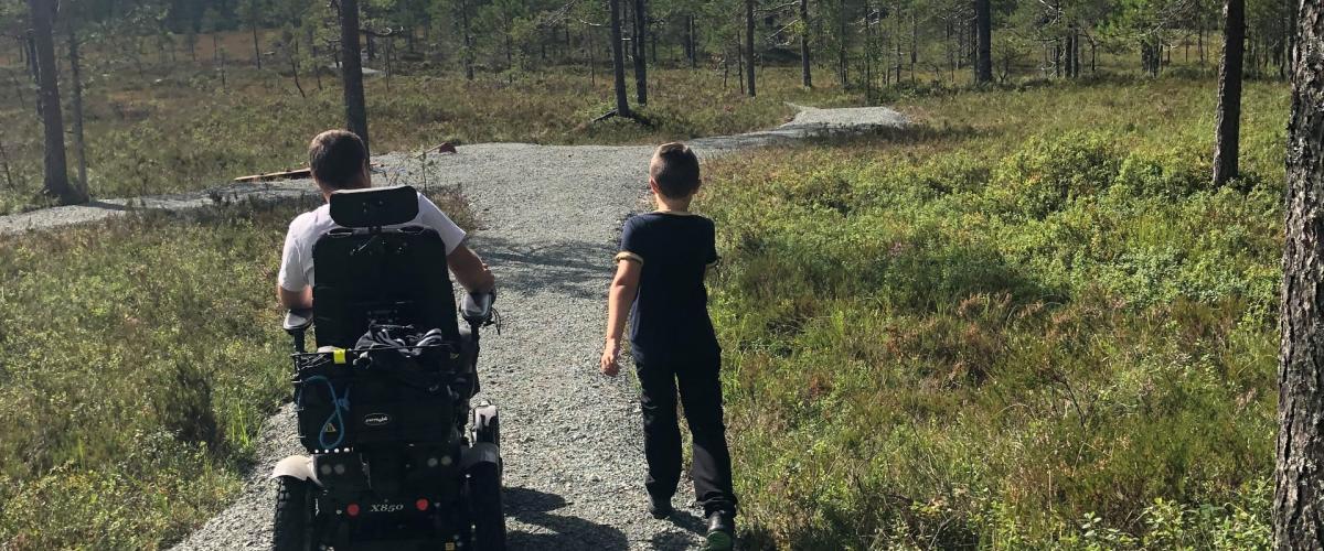 Mann i elektriks rullestol går side om side med et barn på den gruslagte stien