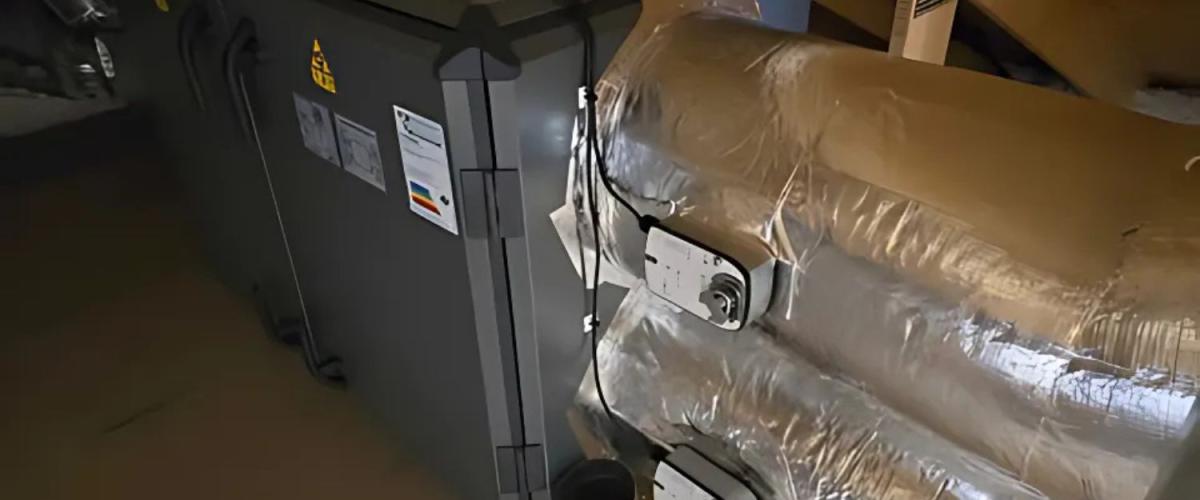 Teknisk rom med ventilasjonsanlegg og isolerte rør. Ventilasjonsanlegget er plassert under et skråtak med synlige kabler og luftkanaler.