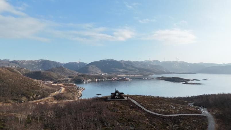 Dagsturhytta ved Utrosjøen ligger på en høyde over fjorden, med utsikt mot øyer, bygda og fjell med vindmøller i bakgrunnen, og en slyngete tursti som leder opp til hytta.