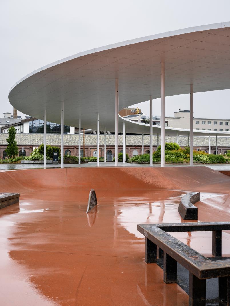 Skateparken på Ruten sett nedenfra, med rødbrune ramper i cortenstål og metallbenker. I bakgrunnen vises gamle murbygninger og den hvite ringen med søyler.