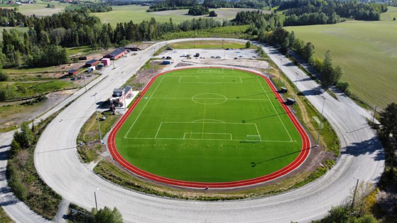 Luftfoto av Bruvollen stadion med kunstgress 11er-bane, rød løpebane rundt og gruslagt omgivelser, omgitt av skog og jordbruksarealer.