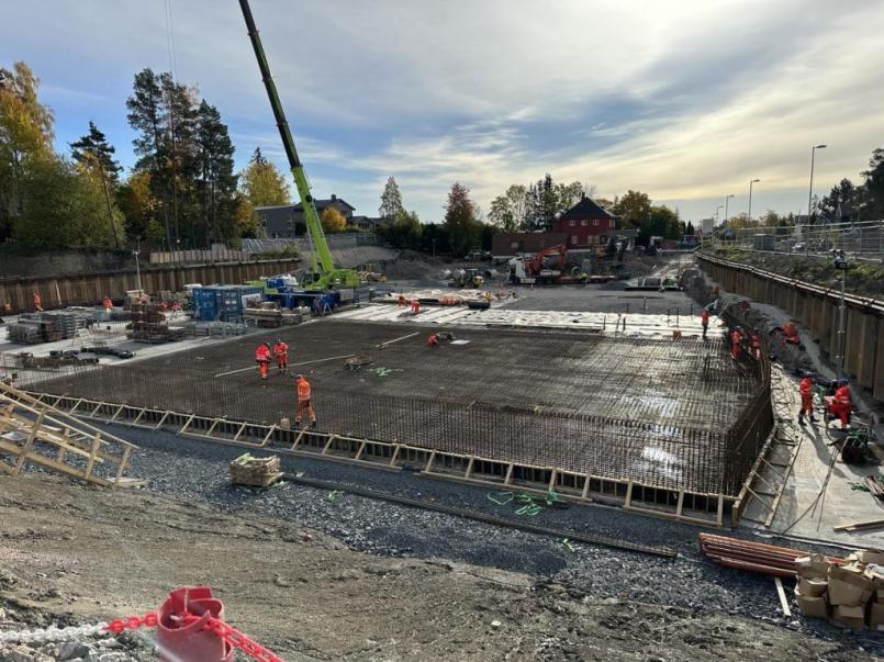 Stor byggeplass på Eckbobanen der arbeidere i oransje refleksklær jobber på en stor armeringsplate for den nye hallen, med kran, anleggsmaskiner og bolighus i bakgrunnen under overskyet himmel.