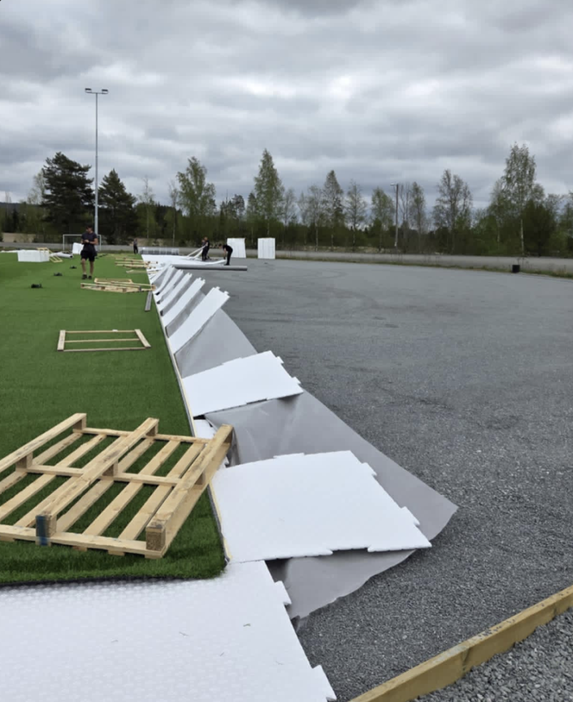 Arbeidere på Bruvollen legger hvite sviktpad-plater langs kanten av kunstgresset, oppå en grå drensduk som ligger på det jevne gruslaget rundt banen.