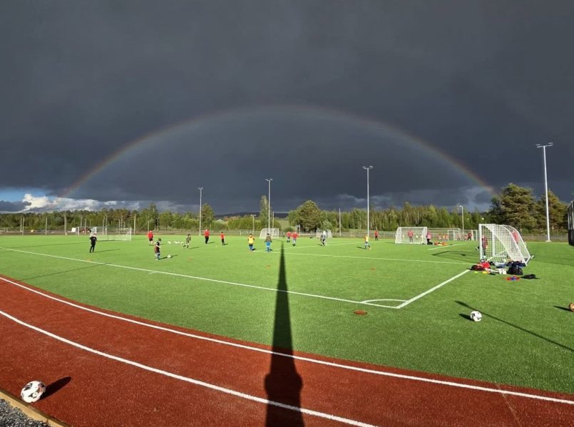 Treningsøkt på kunstgressbanen på Bruvollen under mørke regnskyer, med flere barn som spiller fotball mens en tydelig regnbue spenner seg over banen i bakgrunnen.