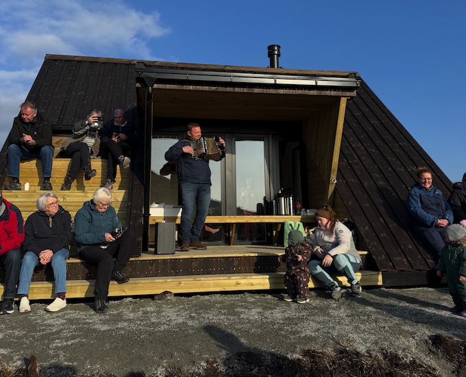 Folk i alle aldre sitter og står utenfor dagsturhytta ved Utrosjøen på åpningsdagen, med kaffe på bordet og en mann som spiller trekkspill på verandaen.