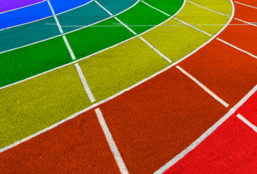 Bildet viser en løpebane på en idrettsplass malt i regnbuens farger. Banen har flere spor i fargene lilla, blå, grønn, gul, oransje og rød, som symboliserer mangfold og inkludering i forbindelse med Pride-bevegelsen. De hvite linjene som skiller sporene er tydelige mot de sterke fargene.